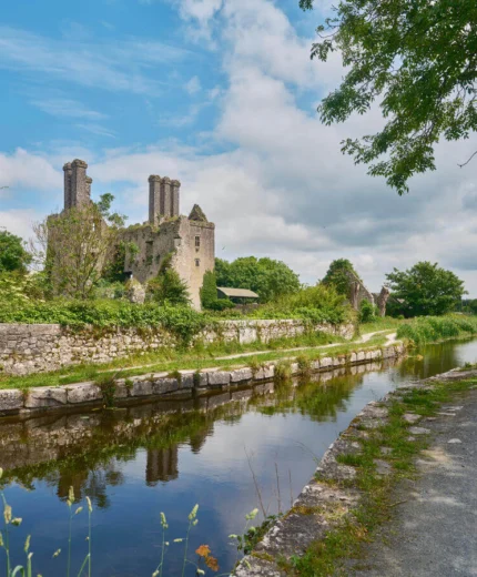 A scenic riverside path by an ancient stone ruin, surrounded by lush greenery under a blue sky.
