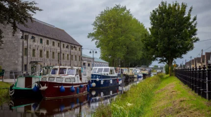 A tranquil canal with moored boats beside an inviting stone building and lush trees.