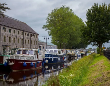 A tranquil canal with moored boats beside an inviting stone building and lush trees.