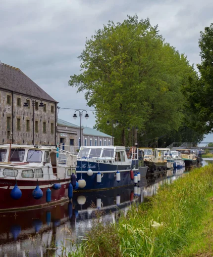 A tranquil canal with moored boats beside an inviting stone building and lush trees.