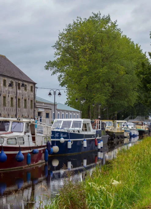 A tranquil canal with moored boats beside an inviting stone building and lush trees.