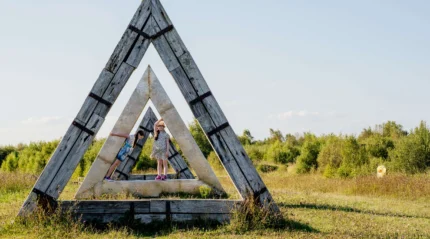 Children playing in large wooden triangle structure in a sunny meadow.