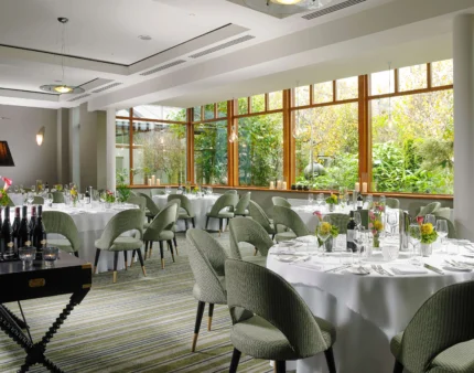 Elegant dining room in a resort with round tables set for guests, large windows revealing lush greenery.