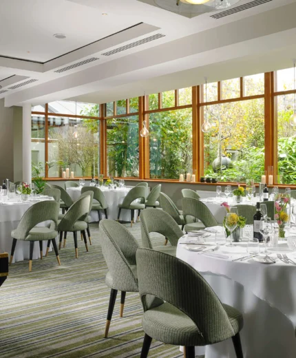 Elegant dining room in a resort with round tables set for guests, large windows revealing lush greenery.