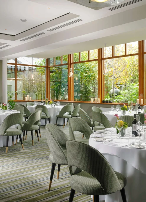 Elegant dining room in a resort with round tables set for guests, large windows revealing lush greenery.