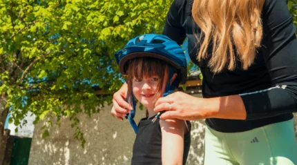 Family outdoors; adult helping child with bike helmet, sunny day.