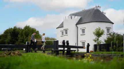 A couple walks along a canal near a white, castle-like rural cottage, under a blue sky.