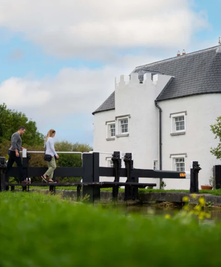 A couple walks along a canal near a white, castle-like rural cottage, under a blue sky.