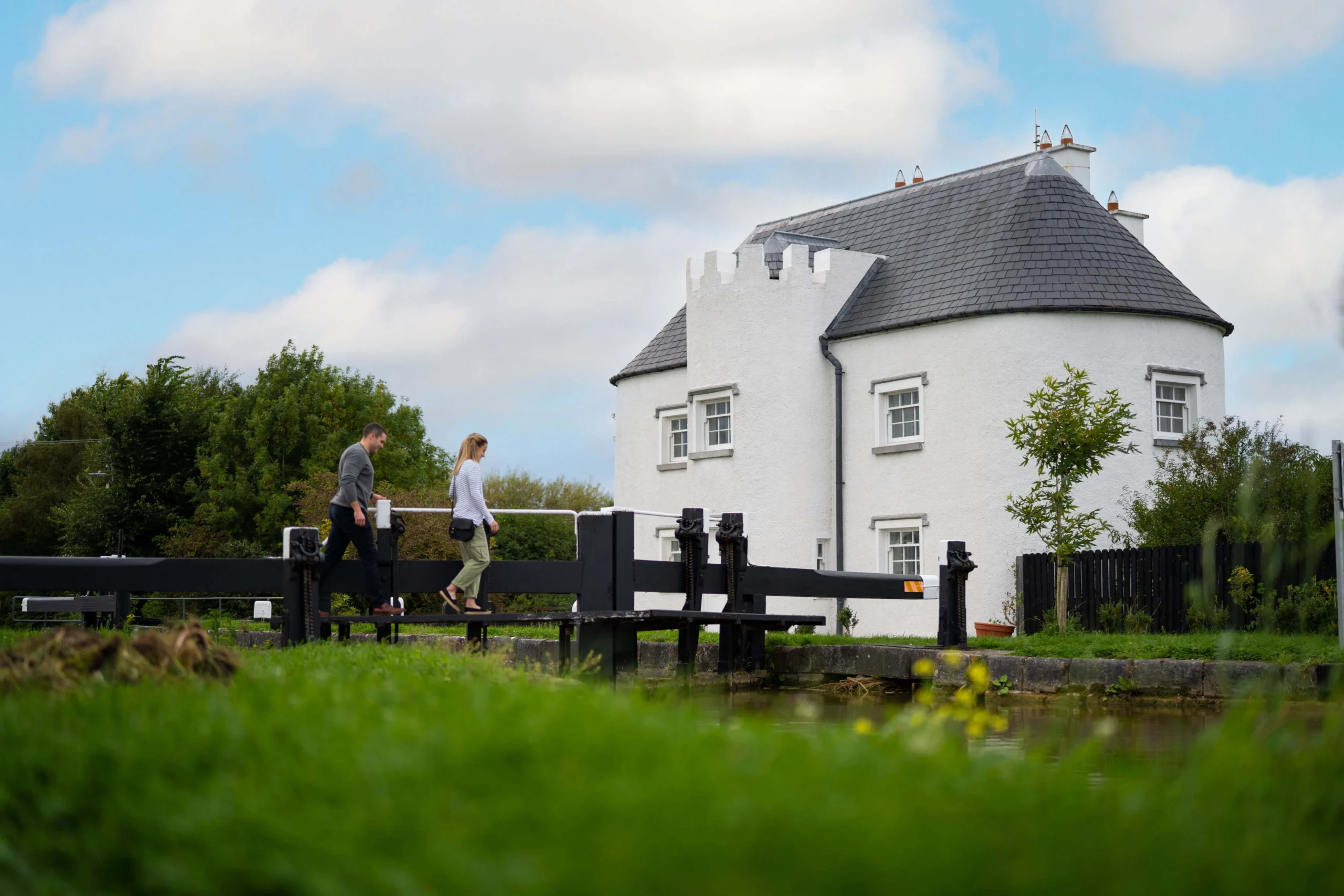 A couple walks along a canal near a white, castle-like rural cottage, under a blue sky.