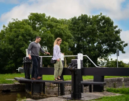 A couple walks hand in hand by a scenic canal in lush greenery.