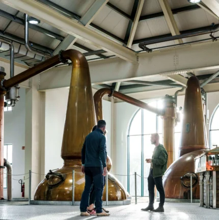 A couple enjoys a guided tour in a distillery with large copper stills in a brightly lit room.