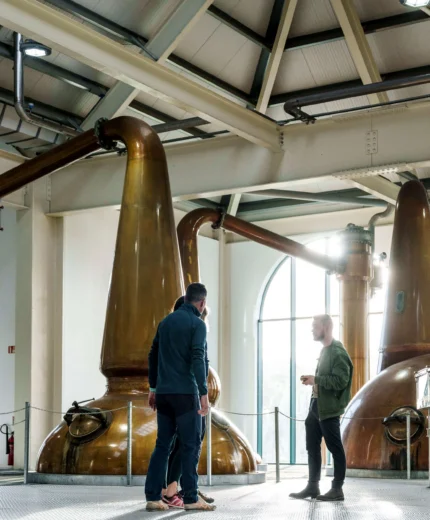 A couple enjoys a guided tour in a distillery with large copper stills in a brightly lit room.