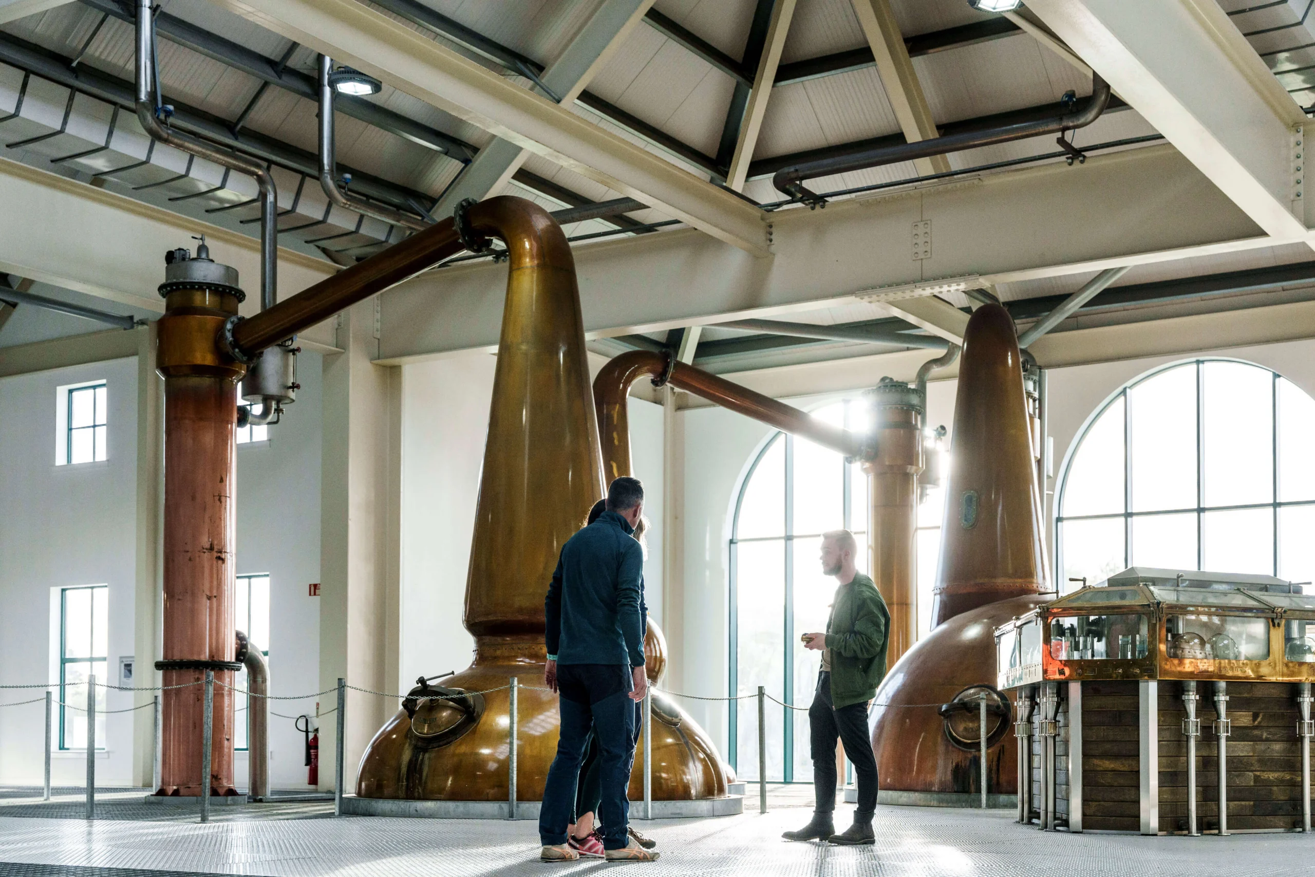 A couple enjoys a guided tour in a distillery with large copper stills in a brightly lit room.