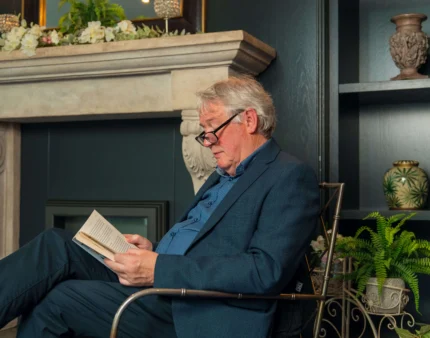 Elderly man reading by a cosy fireplace in a warmly decorated living room.