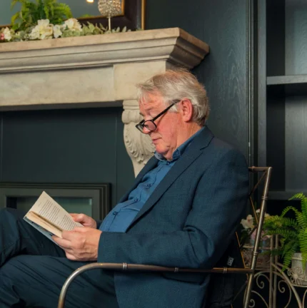 Elderly man reading by a cosy fireplace in a warmly decorated living room.