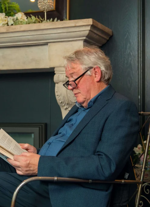 Elderly man reading by a cosy fireplace in a warmly decorated living room.