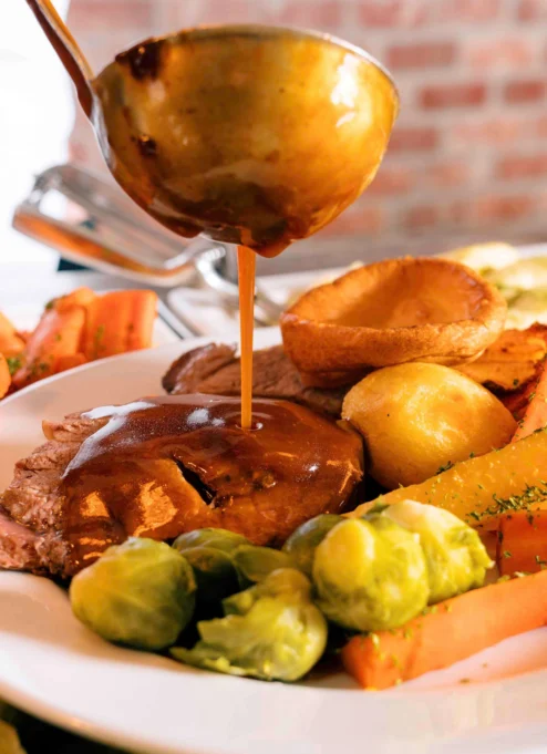 A delicious roast dinner with gravy being poured over tender meat, surrounded by vibrant vegetables.