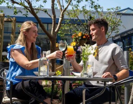 A smiling couple toasts with drinks in a sunny outdoor dining area of a hotel.