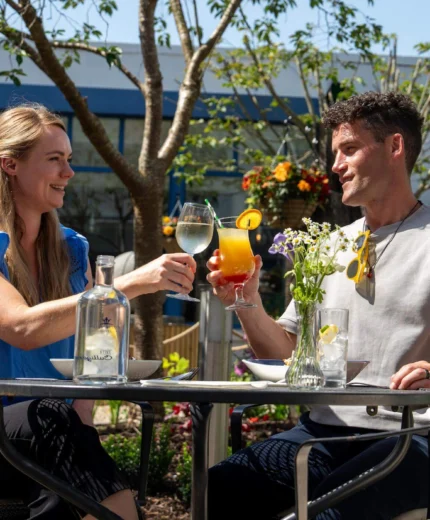 A smiling couple toasts with drinks in a sunny outdoor dining area of a hotel.