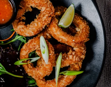 Crispy calamari rings with dip on a dark plate beside fresh greens.