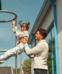 Family enjoying a sunny day as father helps child on playground equipment near resort.