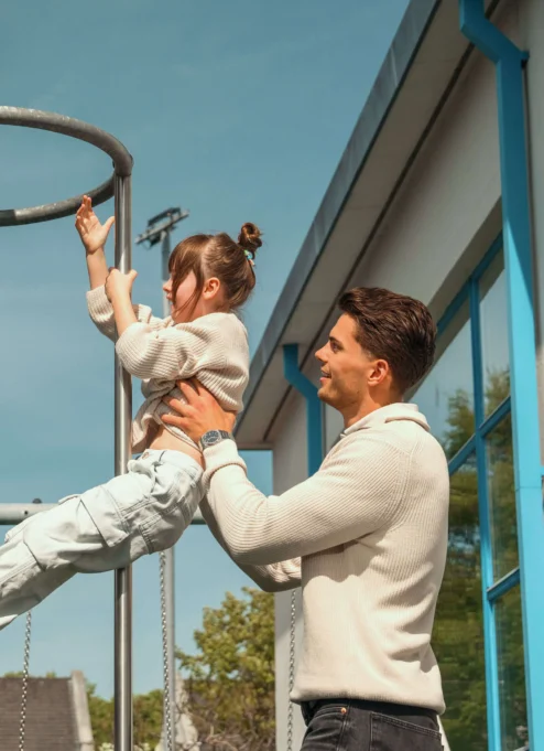 Family enjoying a sunny day as father helps child on playground equipment near resort.
