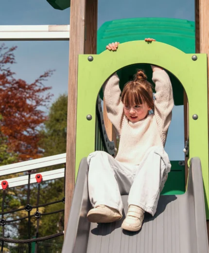 A young girl joyfully slides down a playground slide on a sunny day.