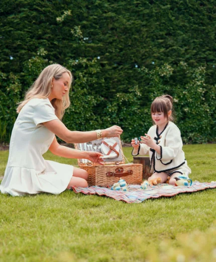 A mother and daughter enjoy a picnic on lush green grass with toys and picnic basket.