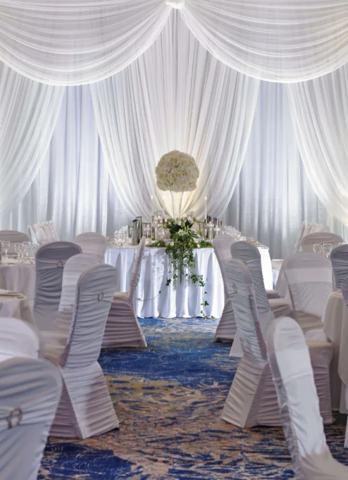 Elegant wedding reception setup with white drapery, round tables, and floral centrepieces.