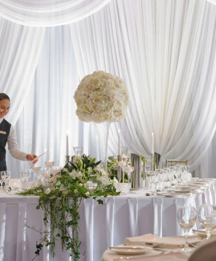 Waitress preparing elegant wedding table in bright, floral-decorated banquet hall.