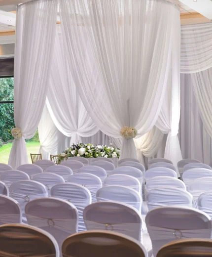 Elegant wedding ceremony setup with white drapes and chairs in a sunlit venue.