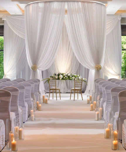 Elegant wedding venue with white drapery, floral arrangements, and candlelit aisle.