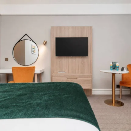 Cosy hotel room with a plush green bedspread, orange chairs, and a flat-screen TV.