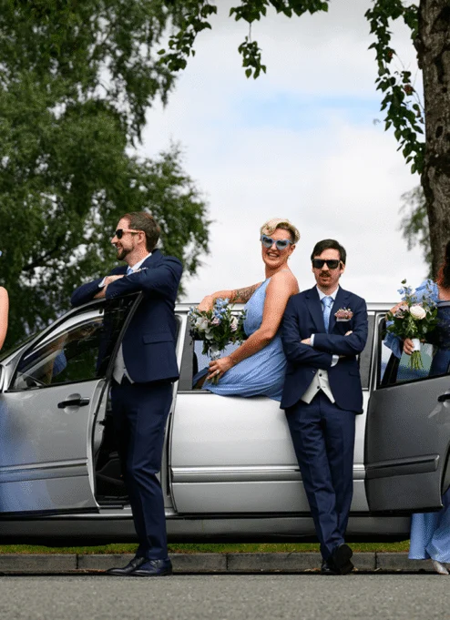 A joyful wedding party in blue outfits posing together by a silver car outside Tullamore Court Hotel.