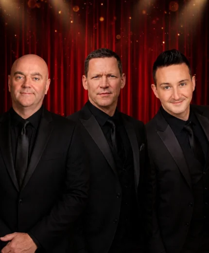 The Three Amigos, three smartly dressed men in black suits smiling on a red theatre curtain backdrop, posing as a friendly performance trio
