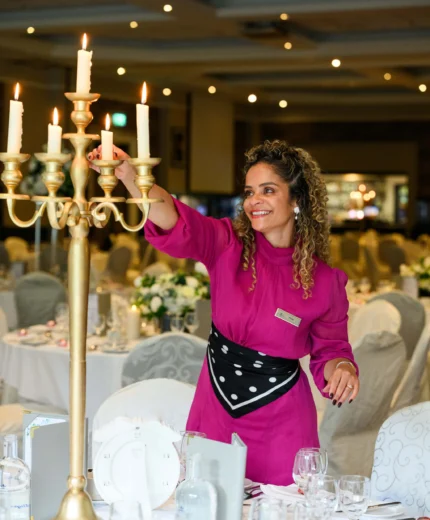 Smiling hotel team member lighting candelabra in a decorated banquet hall, tables set for a family wedding
